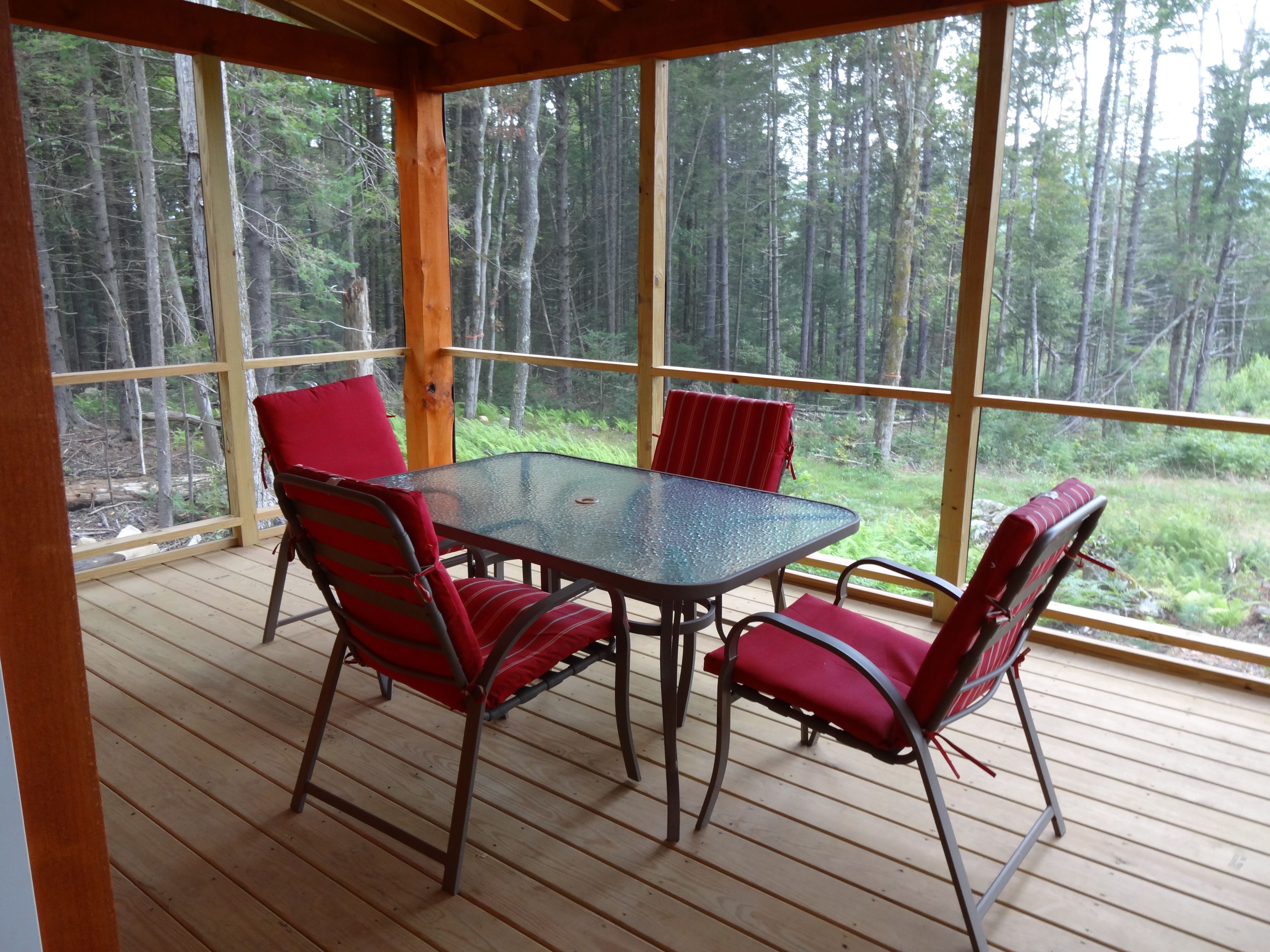 Ett takförsett utomhusområde med ett bord och fyra stolar, omgivet av stora fönster som ger utsikt över en skog.
