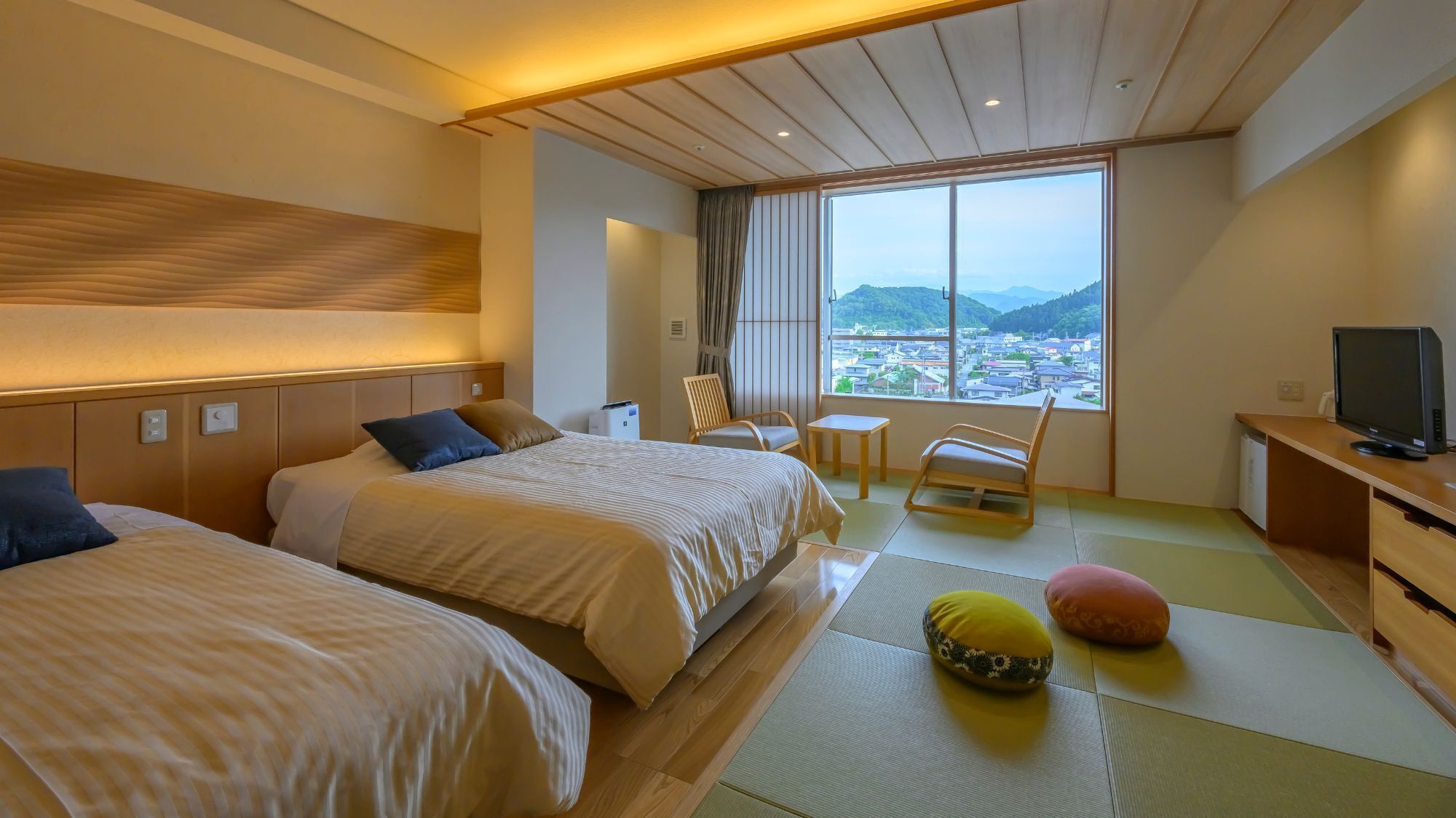 Ein Hotelzimmer mit zwei Betten, einem kleinen Tisch, einem Sessel, einem Fernseher und Blick auf eine Stadt durch große Fenster.