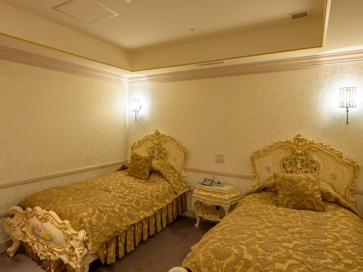 A room with two ornate beds, a small bedside table, and a wall-mounted light fixture.