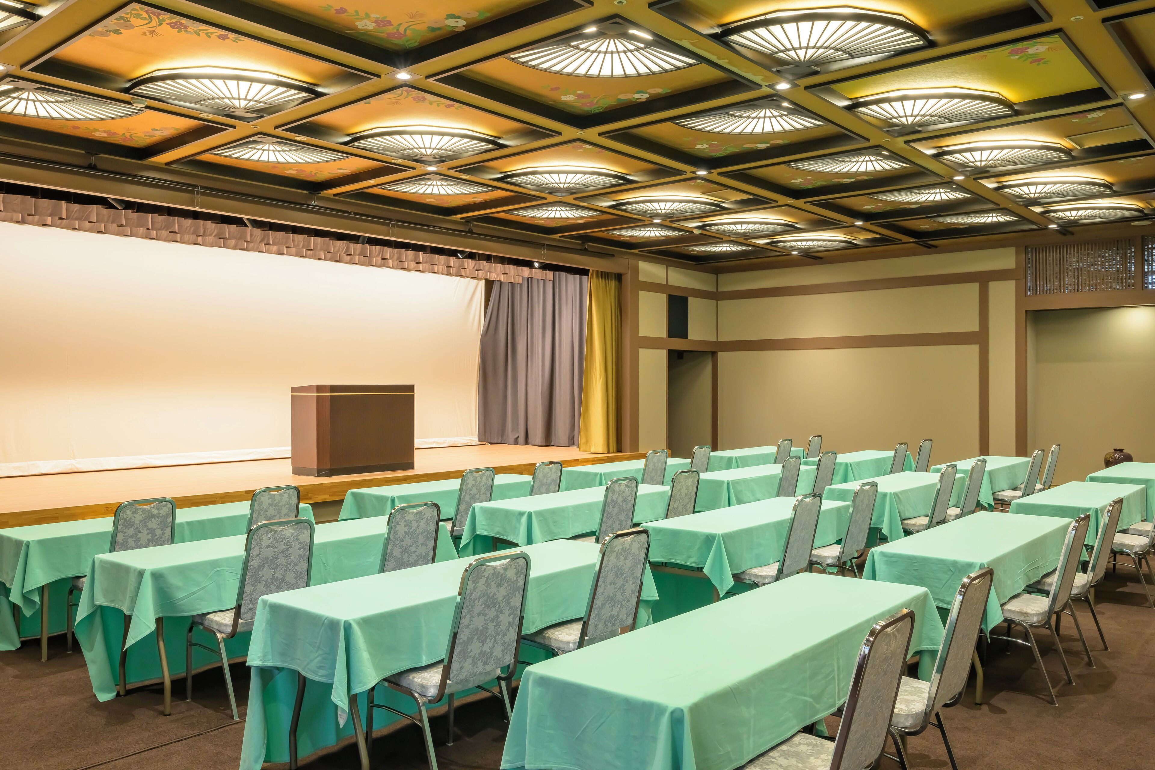 A conference room with rows of tables and chairs, a stage with a podium, and a decorative ceiling.