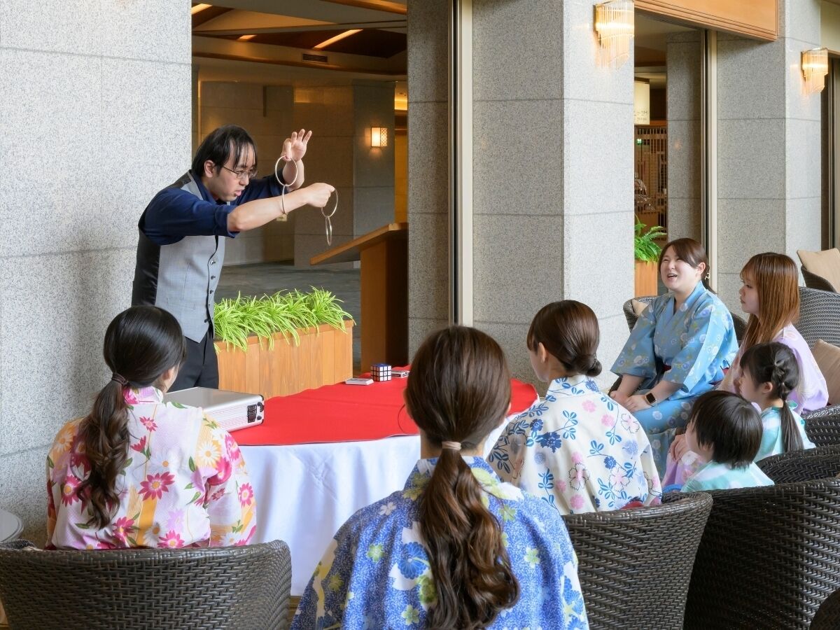A group of people seated outdoors, facing a table with a red cloth and a Rubik's cube.