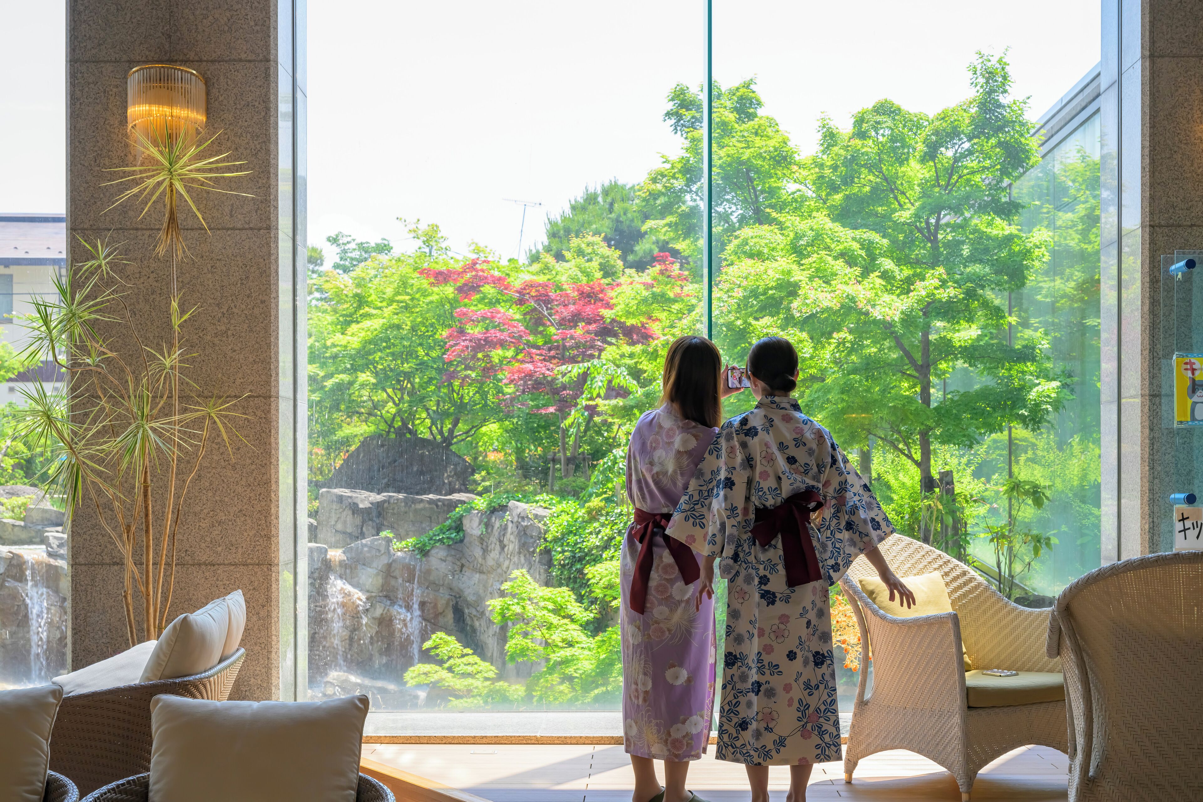 Two people in traditional attire standing by a large window overlooking a waterfall and lush greenery.