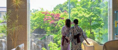 Two people in traditional attire standing by a large window overlooking a waterfall and lush greenery.