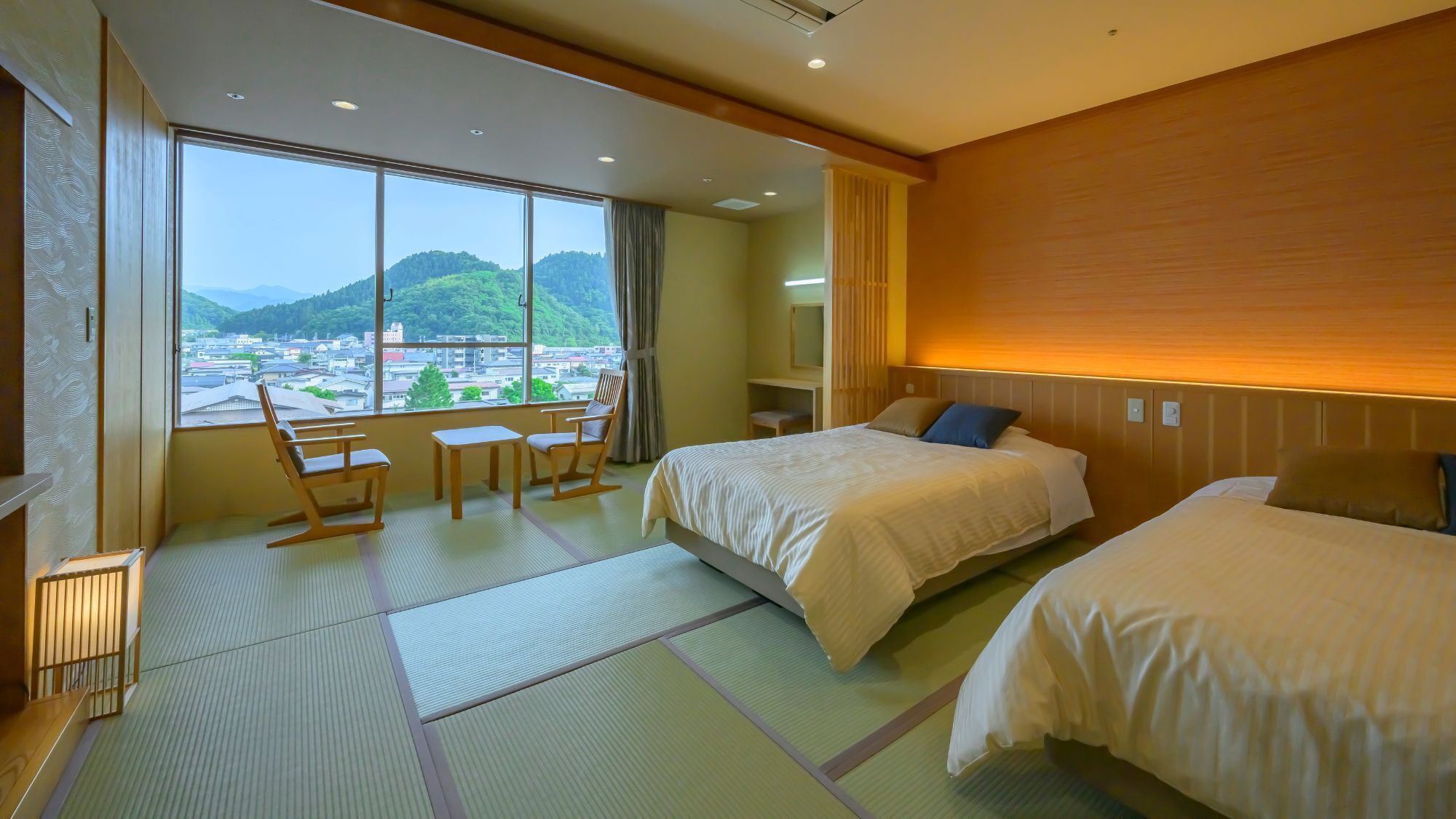 Ein Hotelzimmer mit großem Fenster, zwei Betten, einer Sitzecke mit Sesseln und Blick auf Stadt und Berge.