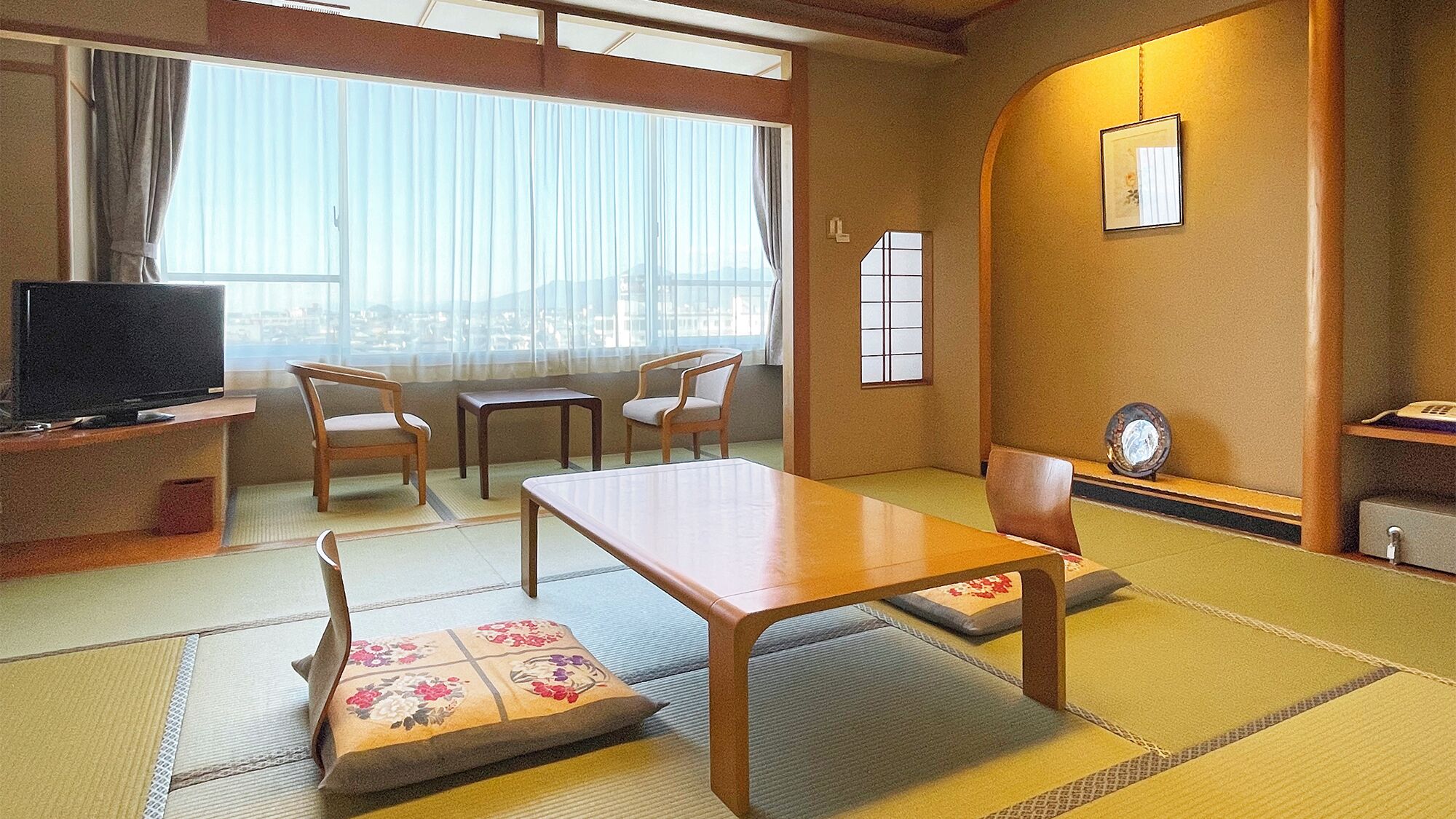 Ein traditioneller japanischer Raum mit Tatami-Böden, einem niedrigen Tisch, zwei Stühlen, einem Fernseher und einer Wanduhr.