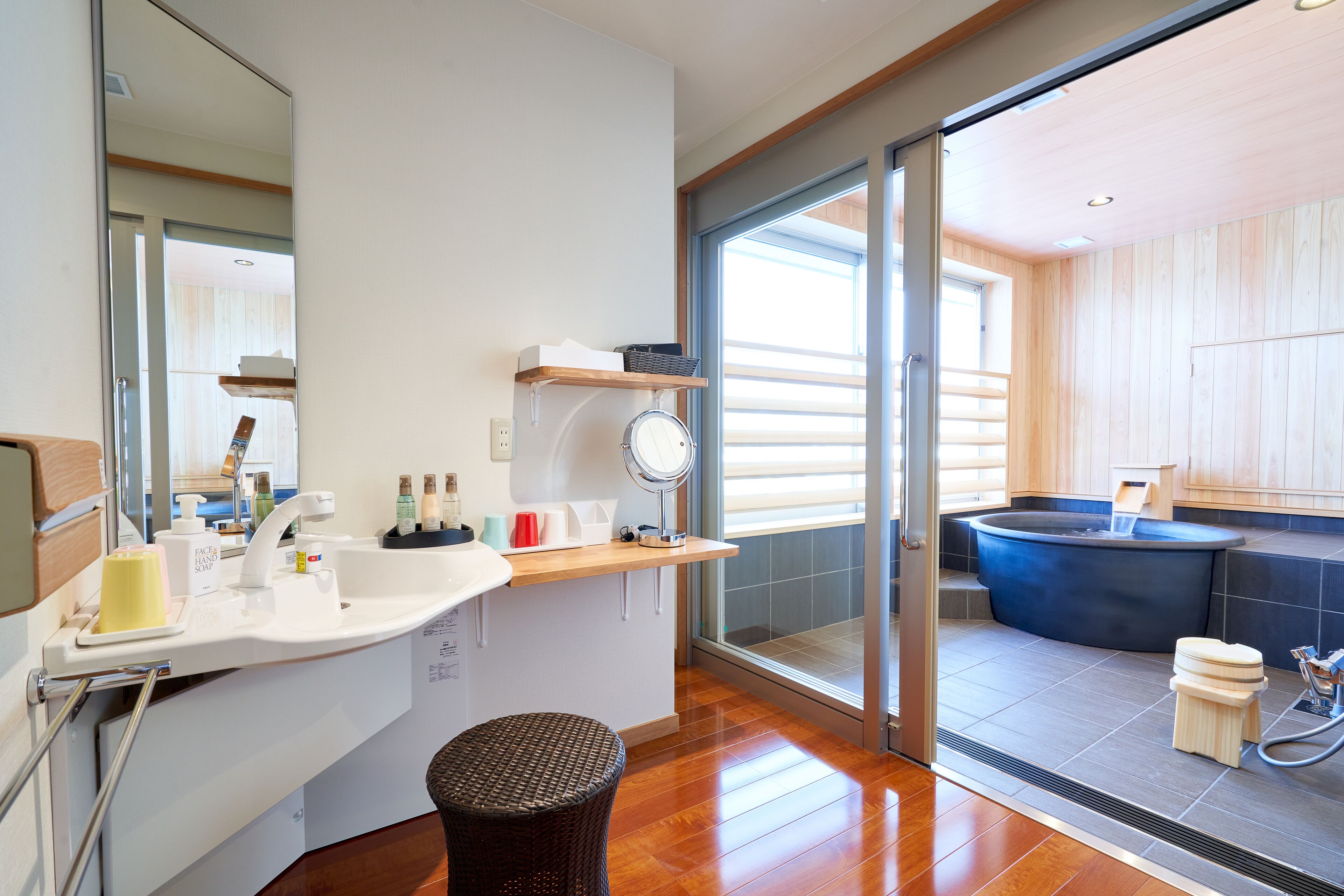 Un baño moderno con un espejo grande, un lavamanos con artículos de aseo, un estante de madera y una bañera de hidromasaje.