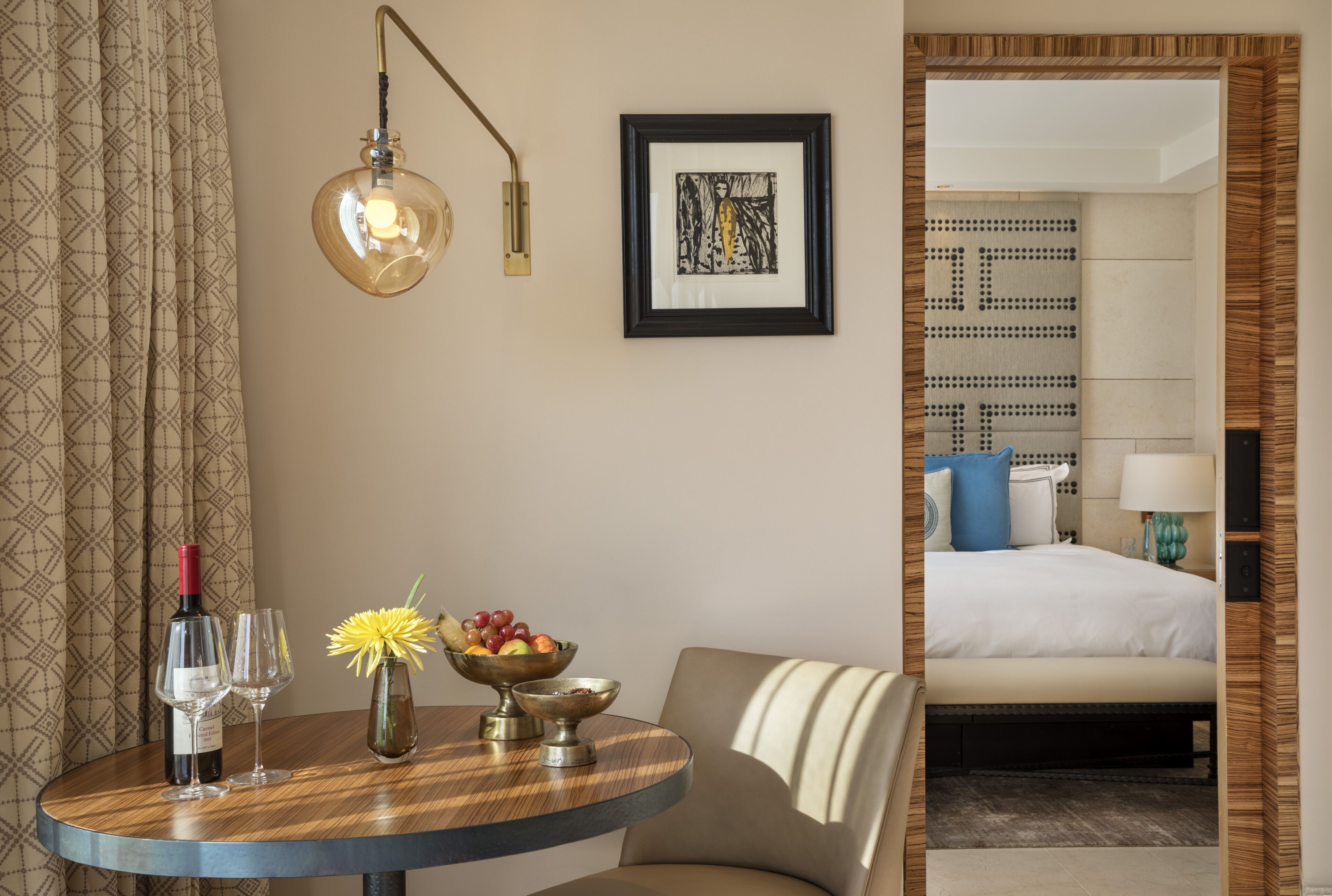 A hotel room with a bed, a bedside table with a wine bottle, glasses, and fruit, a framed picture on the wall, and a modern lamp.