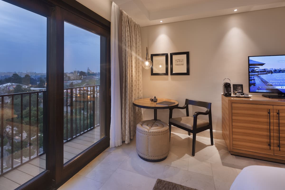 Habitación de hotel con un ventanal grande, una mesa redonda, una silla, un pequeño reposapiés, un armario y un televisor.