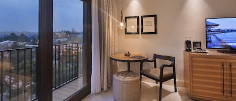 Habitación de hotel con un ventanal grande, una mesa redonda, una silla, un pequeño reposapiés, un armario y un televisor.