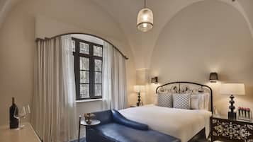 Habitación de hotel con una cama grande, una mesita con una botella y vasos, un sillón azul y una mesita de noche con lámpara.