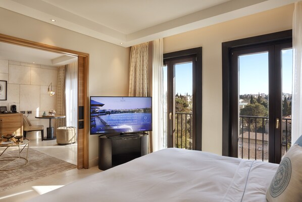 A hotel room with a bed, a TV, a balcony with a city view, and a dining area with a table and chairs.