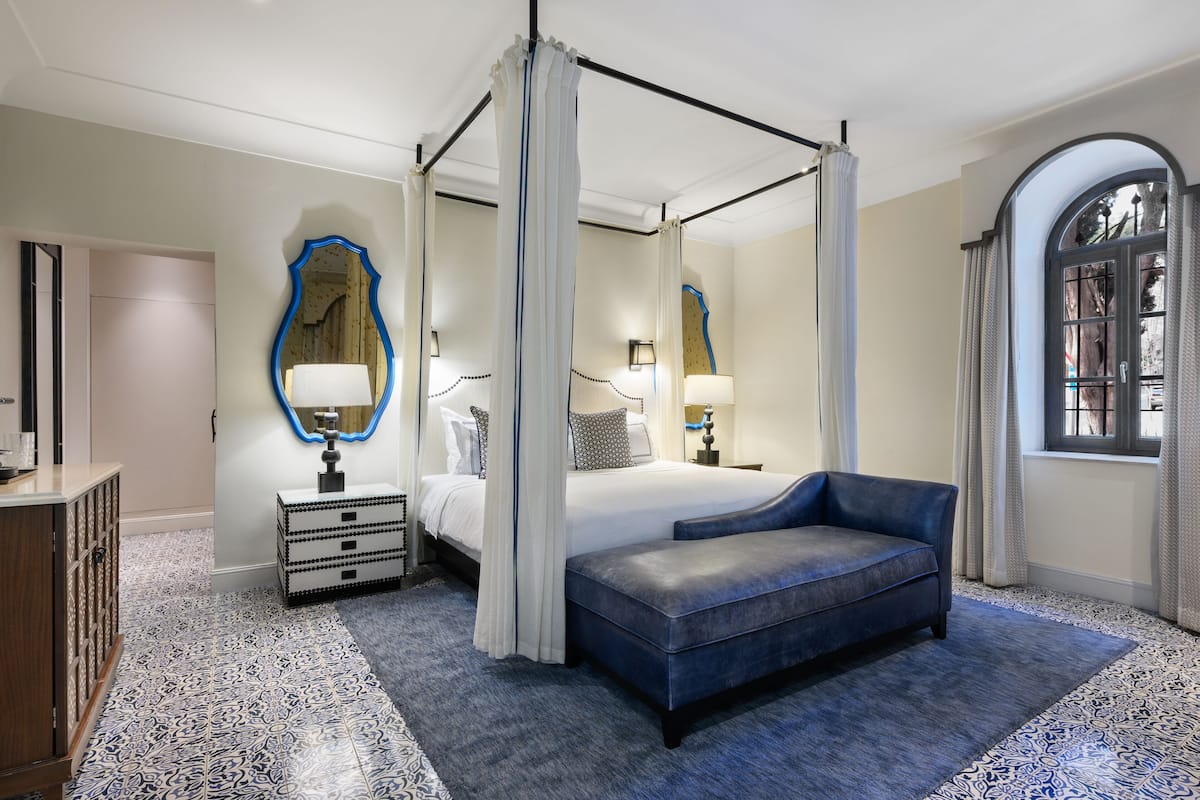 Una cama con dosel y cuatro columnas, un banco tapizado en azul, un espejo grande con marco azul y un suelo estampado.