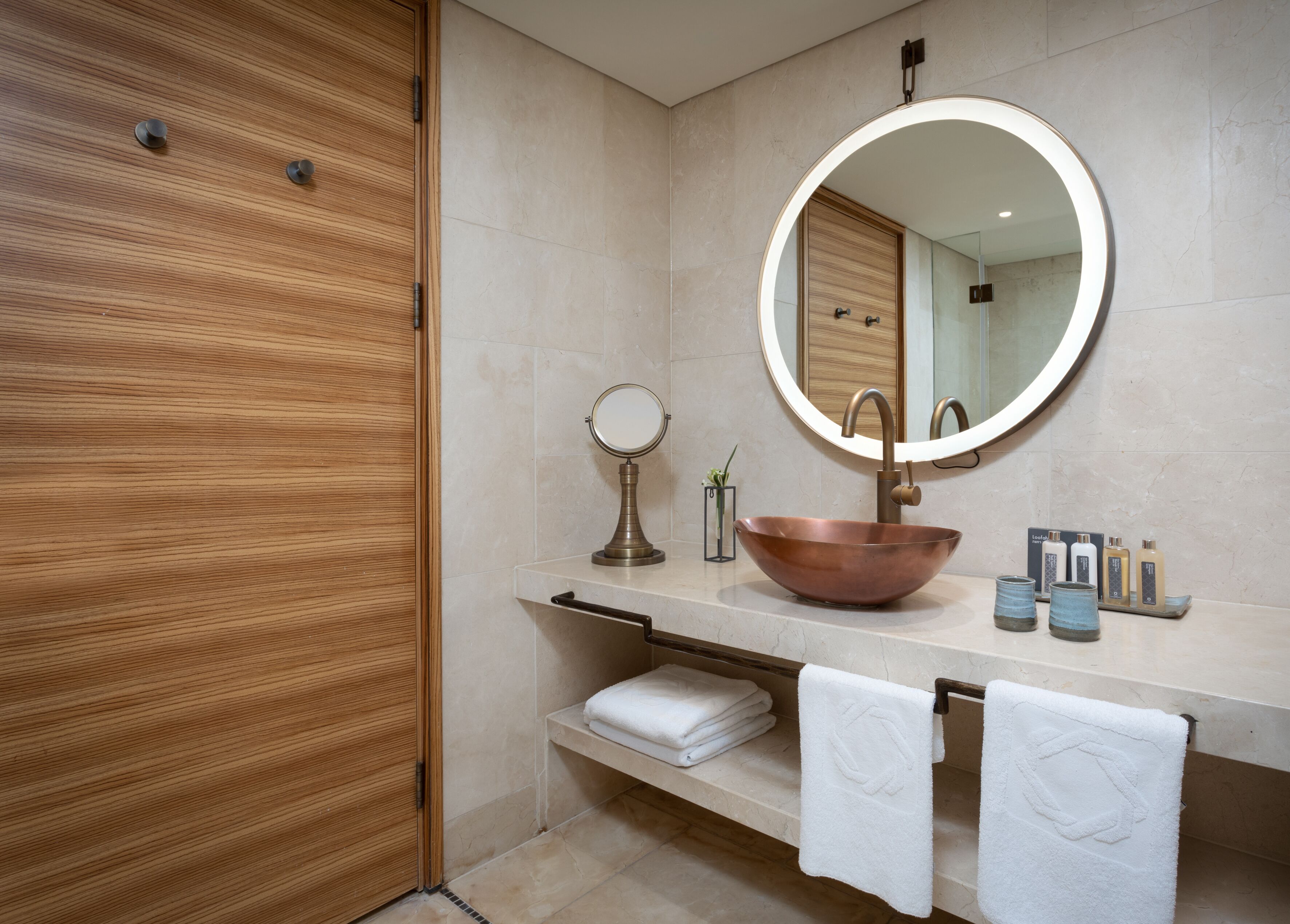 A modern bathroom with a wooden door, a round mirror, a copper sink, and neatly folded towels.