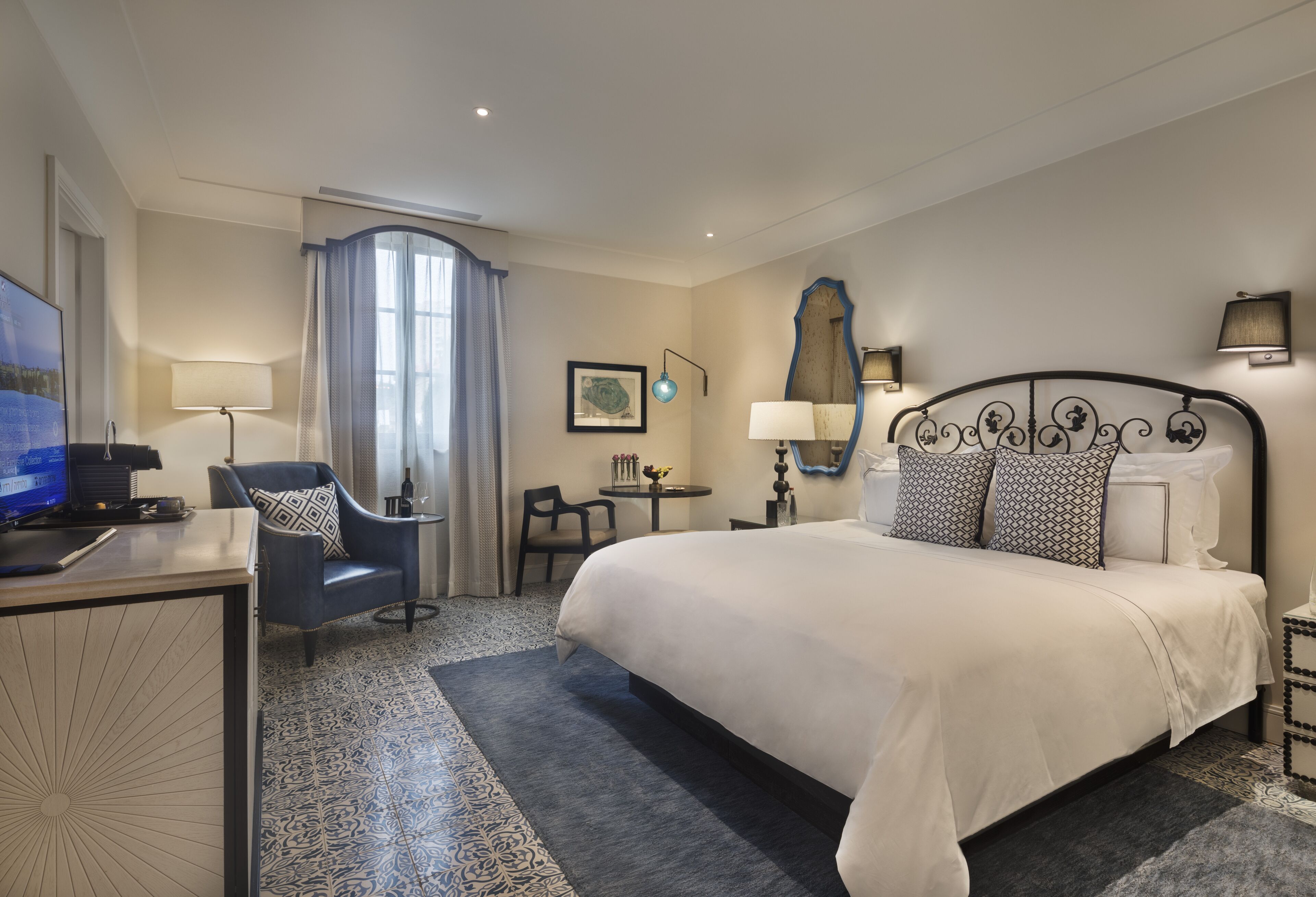 A hotel room with a large bed, a desk with a computer, a chair, a small table, and a mirror.