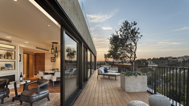 A modern balcony with wooden flooring, a seating area with a sofa and chairs, and a view of the cityscape. - Orient by Isrotel exclusive (Jerusalem)