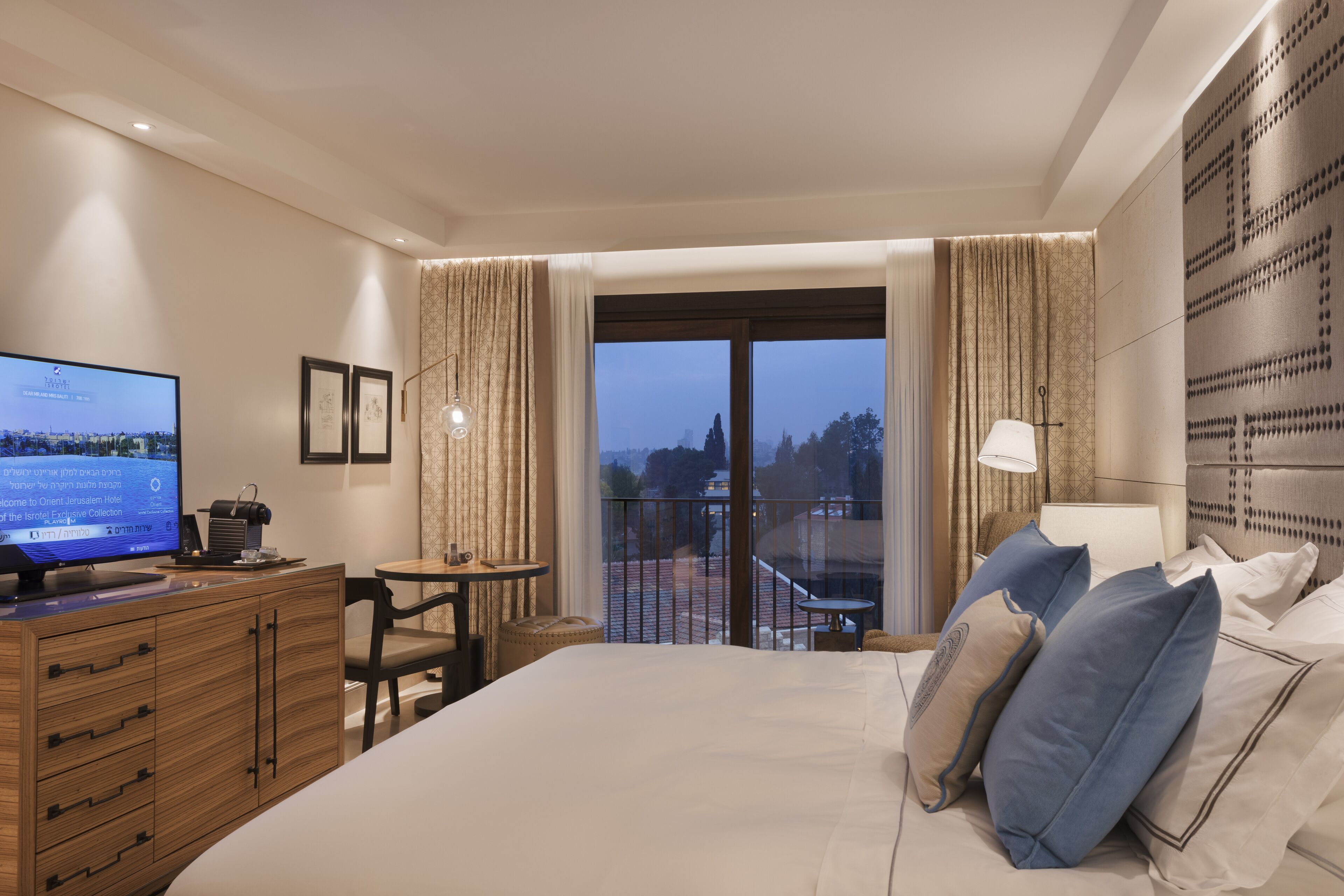 Habitación de hotel con cama, escritorio, televisor y balcón con cortinas.