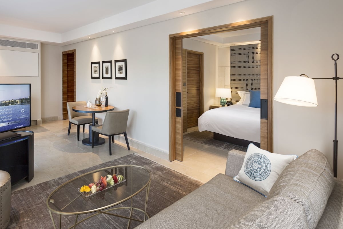 Habitación de hotel con cama, zona de estar con mesa y sillas, televisión y mesa de centro con fruta.