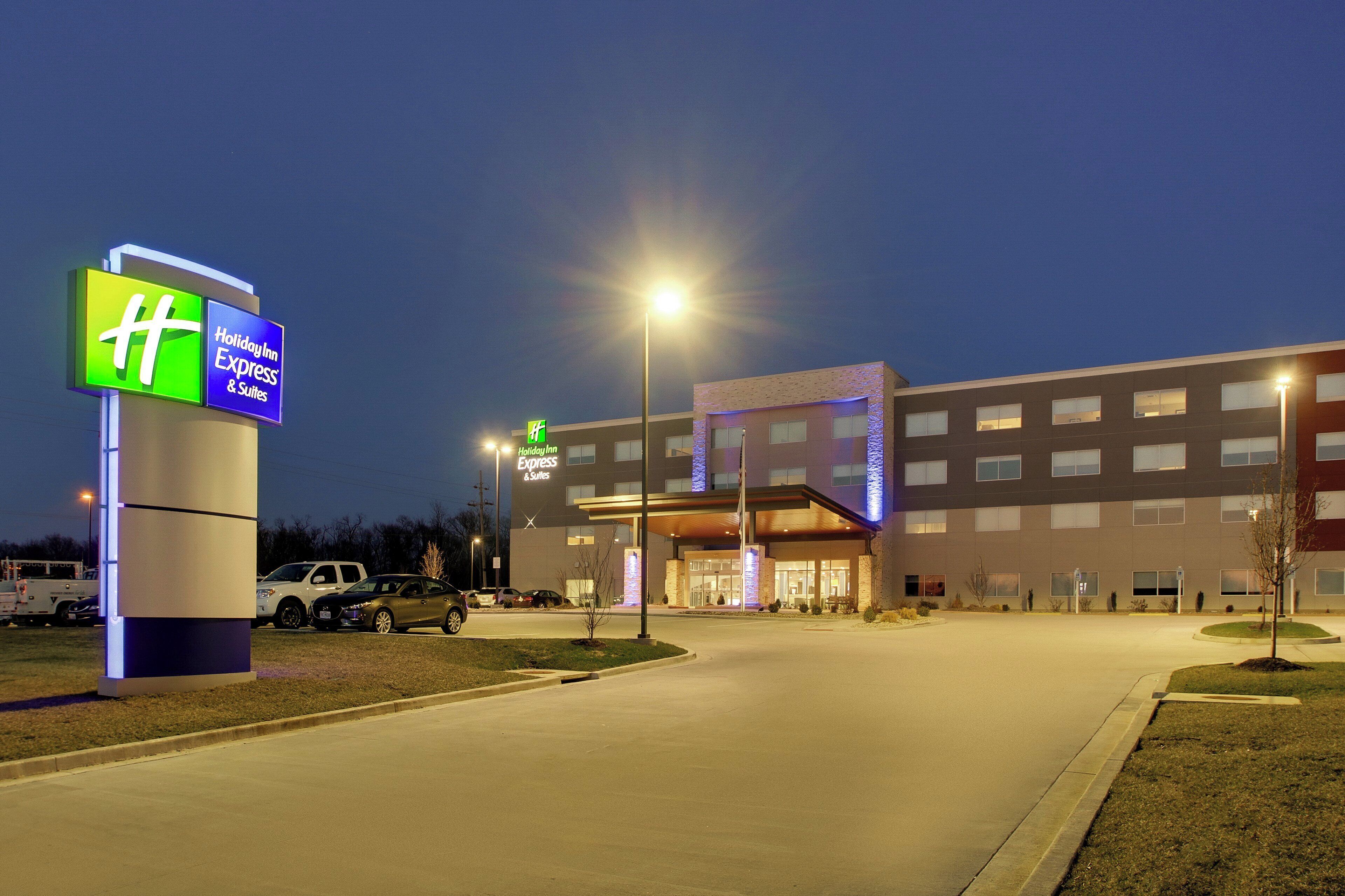 Un hotel Holiday Inn Express & Suites amb una façana de disseny modern, una entrada ben il·luminada i una zona d'aparcament amb diversos cotxes.