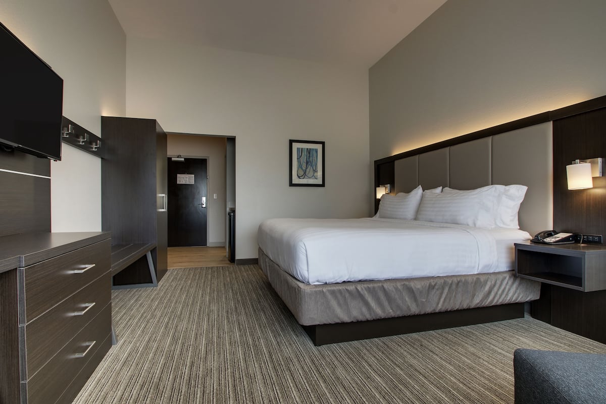 A modern hotel room with a large bed, a desk with drawers, a flat-screen TV, and a bedside table with a lamp.