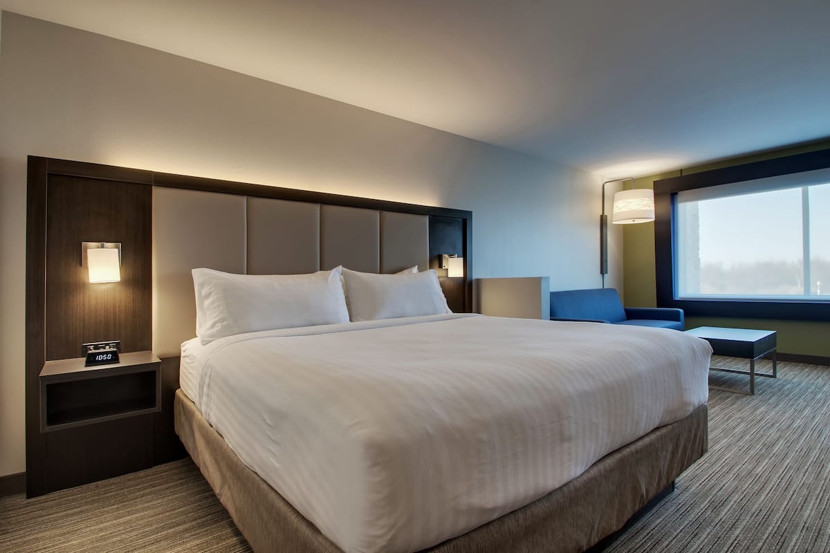 A hotel room with a large bed, a nightstand with a clock, a bedside table, a chair, and a large window.
