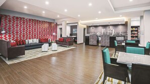 A modern hotel lobby with a patterned wall, a seating area with sofas and chairs, a dining area with tables, and a kitchen in the background. - La Quinta by Wyndham San Antonio by Frost Bank Center (San Antonio)