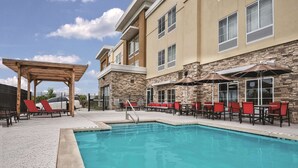 A pool area with lounge chairs and umbrellas, a pergola, and a stone building with multiple windows. - La Quinta by Wyndham San Antonio by Frost Bank Center (San Antonio)