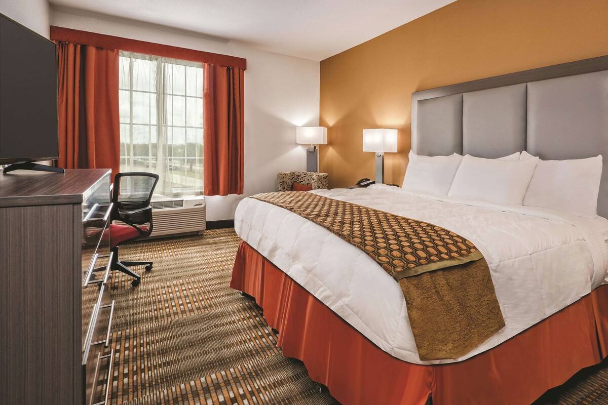A hotel room with a bed, desk, chair, lamp, and window with curtains.
