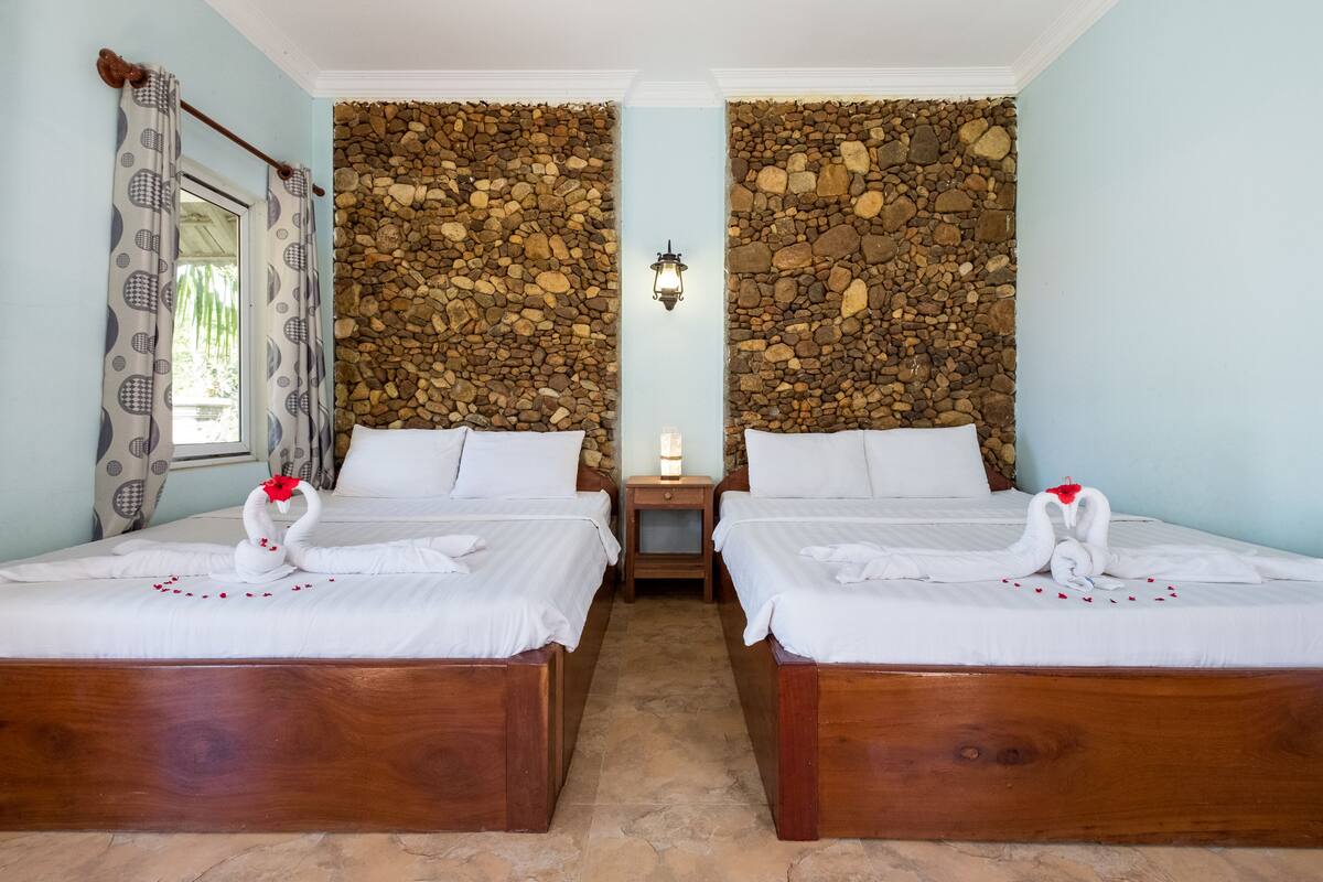A room with two wooden beds, a stone wall, a nightstand with a lamp, and a window with patterned curtains.