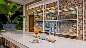 Zona de bar con pared de piedra, estantes con botellas y copas, una planta en maceta y un plato de galletas.