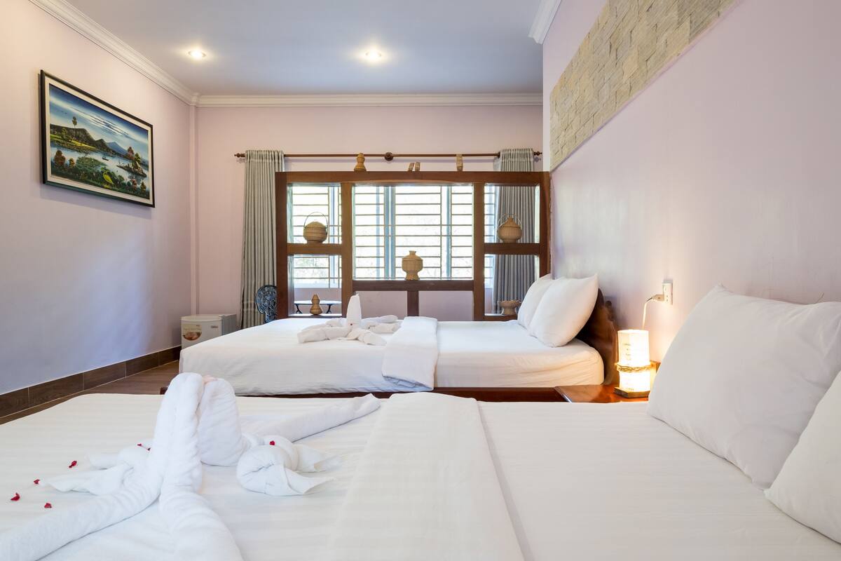 A hotel room with two beds, a window with blinds, a bedside table with a lamp, and a painting on the wall.