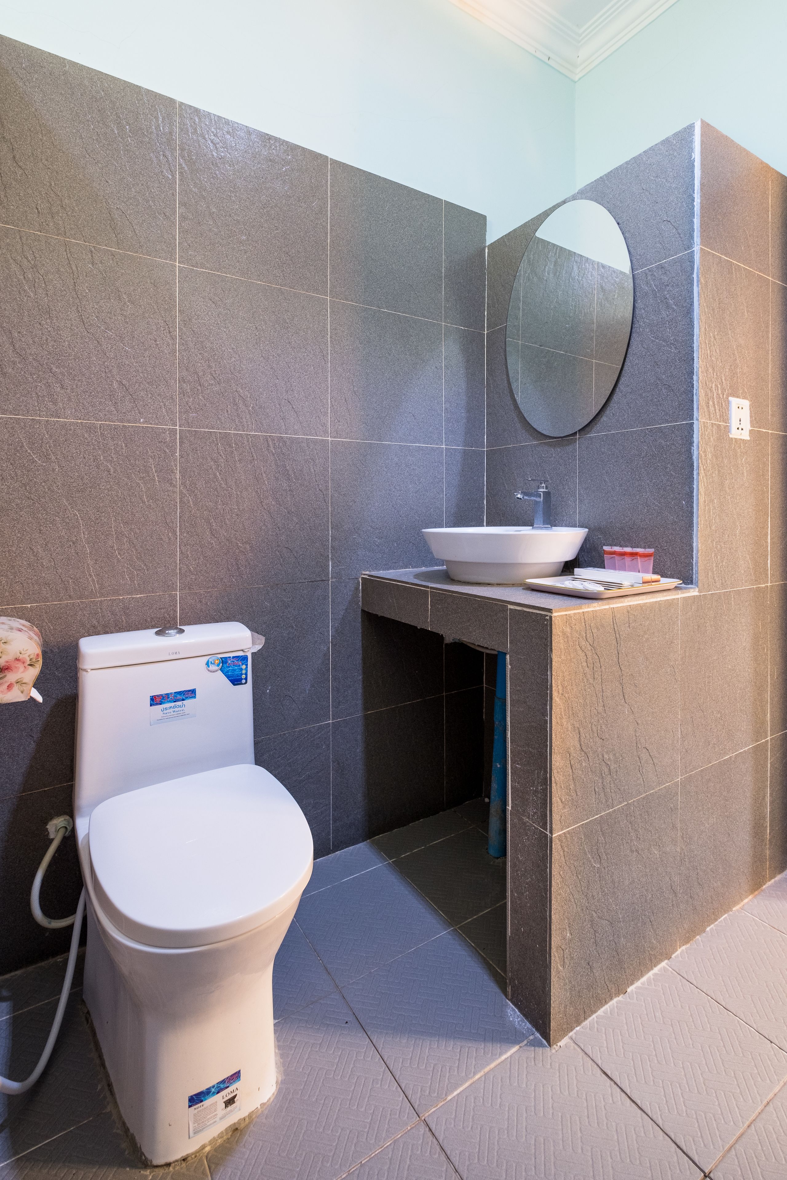 Un baño moderno con inodoro blanco, lavamanos con espejo y paredes de baldosas oscuras.