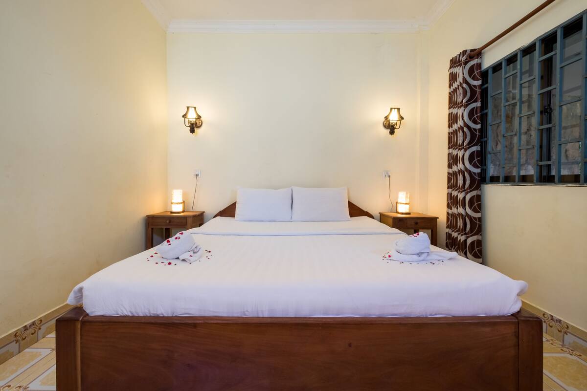 A neatly made bed with white linens and pillows, two bedside lamps, and a window with patterned curtains.