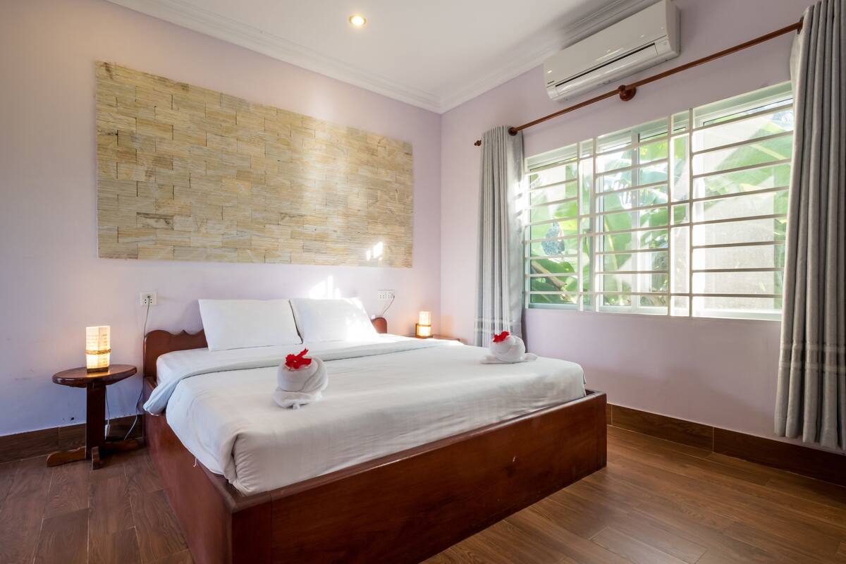 A bedroom with a wooden bed, a wall-mounted air conditioner, a bedside table with a lamp, and a window with curtains.