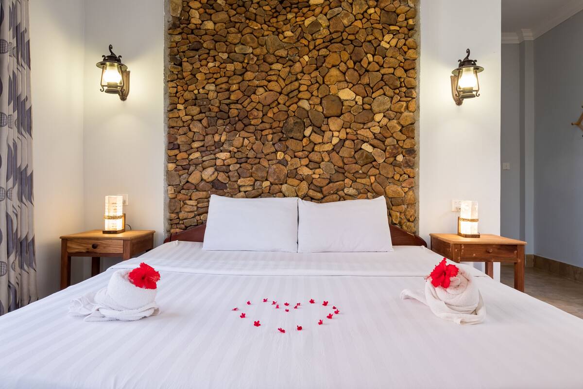 A neatly made bed with white linens, a heart-shaped arrangement of red flowers on the bed, and two towels with red flowers on the ends.
