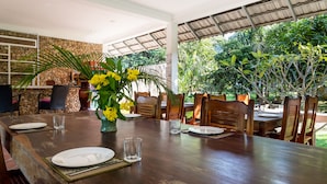 Comedor con una larga mesa de madera dispuesta para cuatro personas, rodeada de sillas, una pared de piedra y vistas al exterior con vegetación.