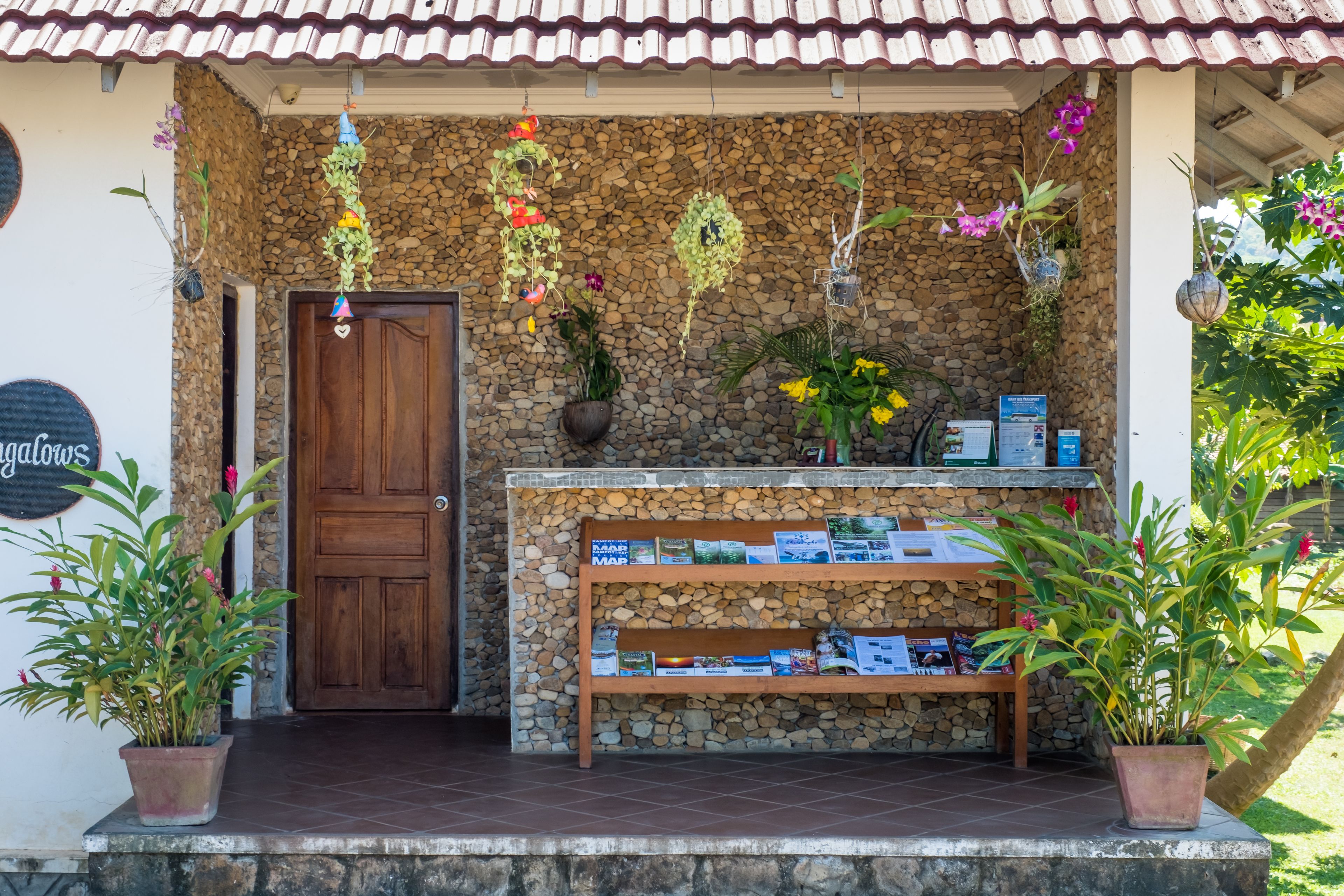 A rustic building with a stone wall, a wooden door, and a shelf with books and plants.