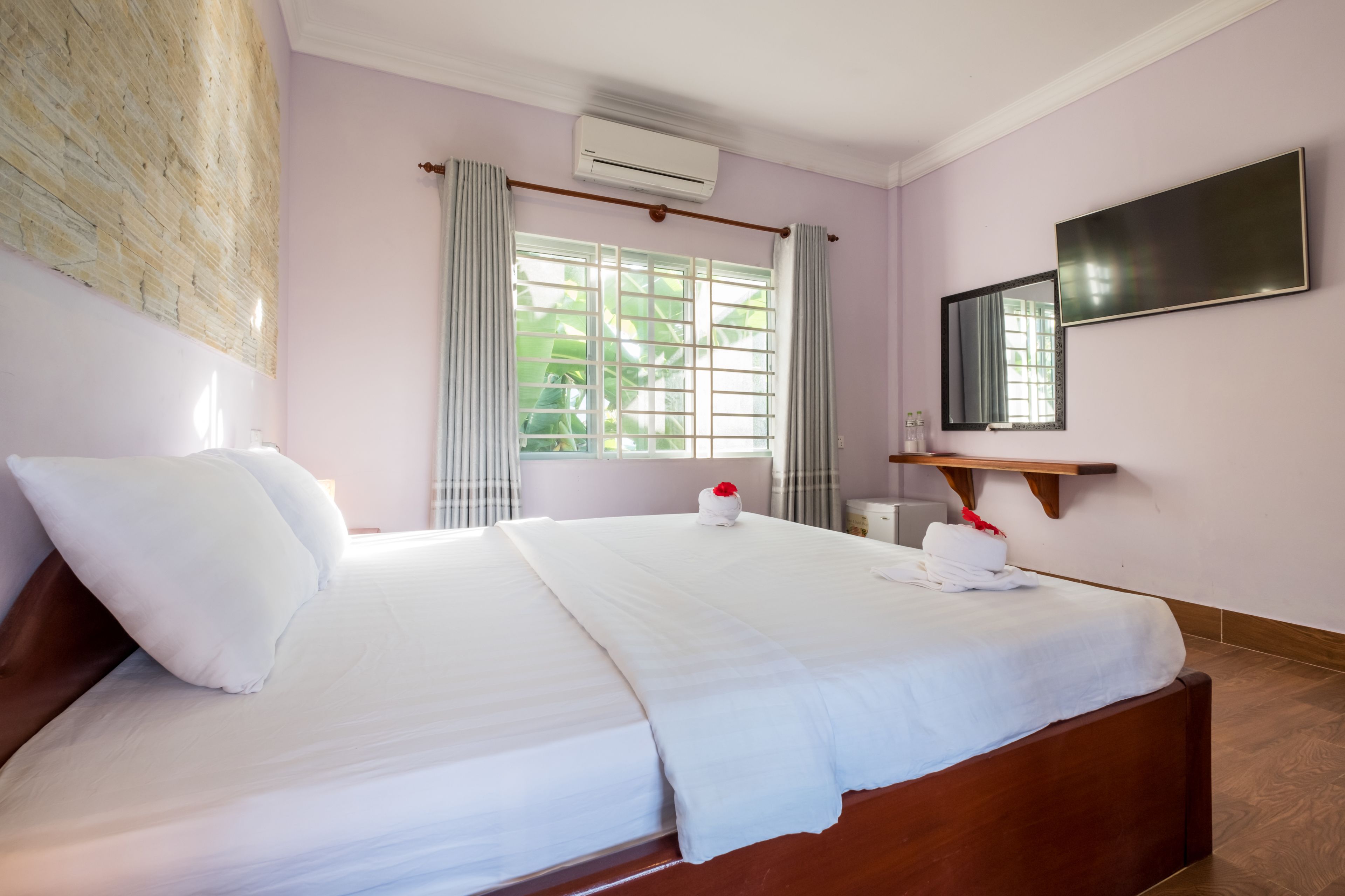 A neatly made bed with white linens and pillows, a wooden headboard, a bedside table with a mirror, and a window with curtains.