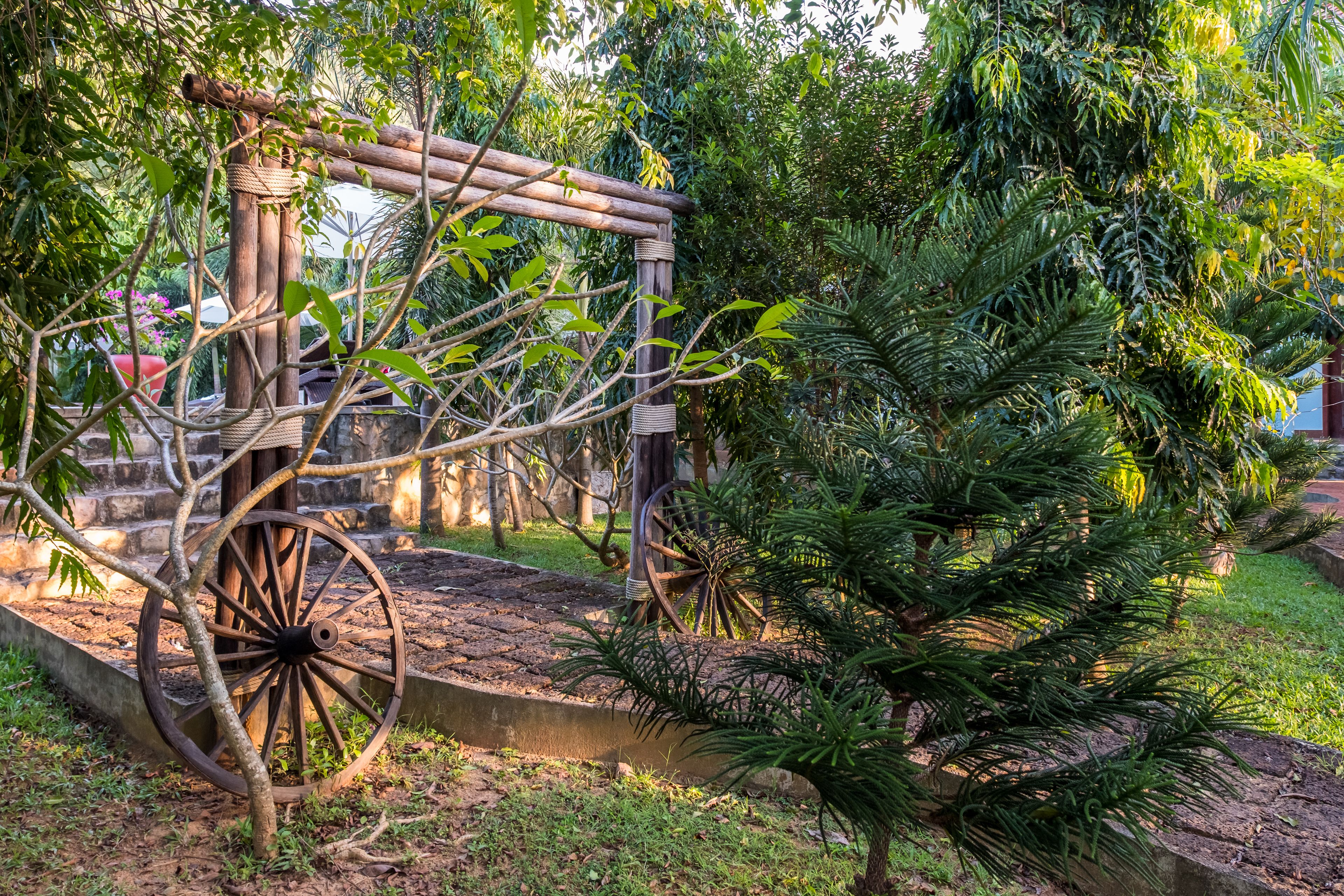 A rustic wooden structure with a wheel, surrounded by lush greenery and a concrete pathway.