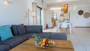 Un saló modern amb un sofà gris, una taula de centre de fusta amb un plat de fruita, una zona de menjador amb una taula i cadires blanques, i una cuina al fons.