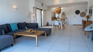 Un saló modern amb un sofà gris, una taula de centre de fusta amb fruita, una zona de menjador amb taula i cadires blanques, i una cuina al fons.