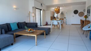 A modern living room with a grey sofa, a wooden coffee table with fruit, a dining area with a white table and chairs, and a kitchen in the background.