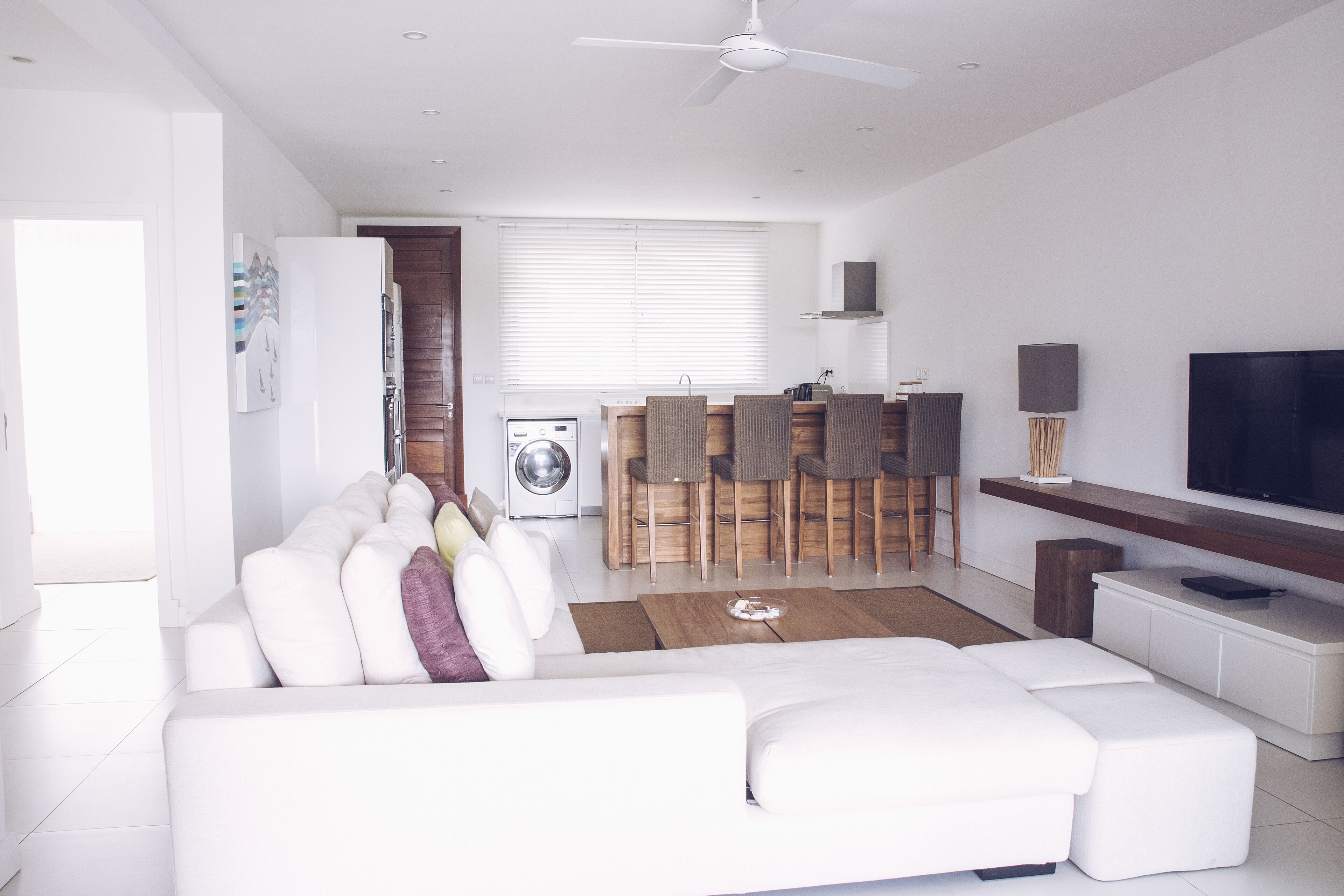 A modern living room with a white sofa, a dining area with wooden chairs, and a TV on a wooden shelf.