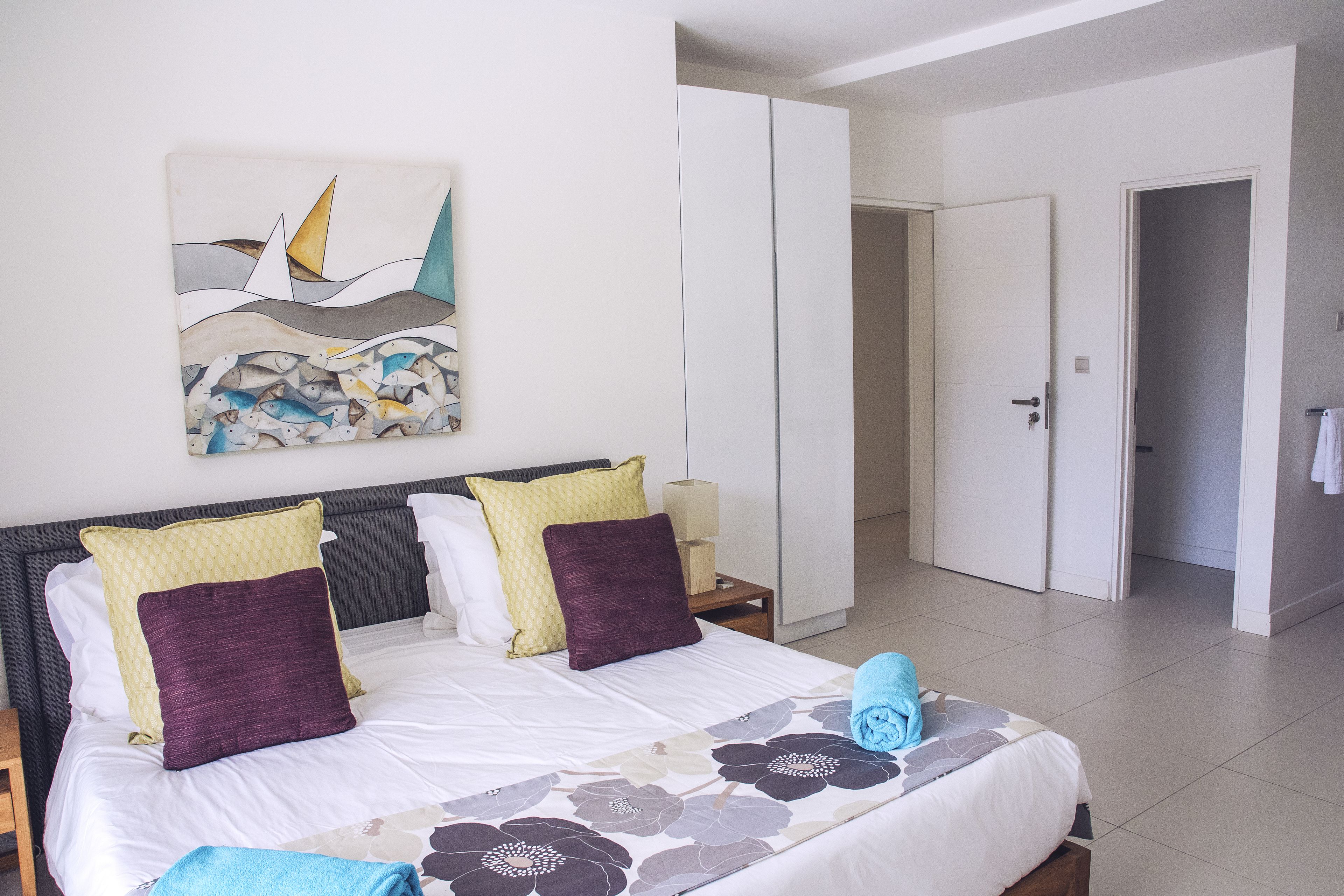 A neatly made bed with a floral bedspread, two pillows, and a blue towel. A painting of sailboats and fish hangs on the wall.