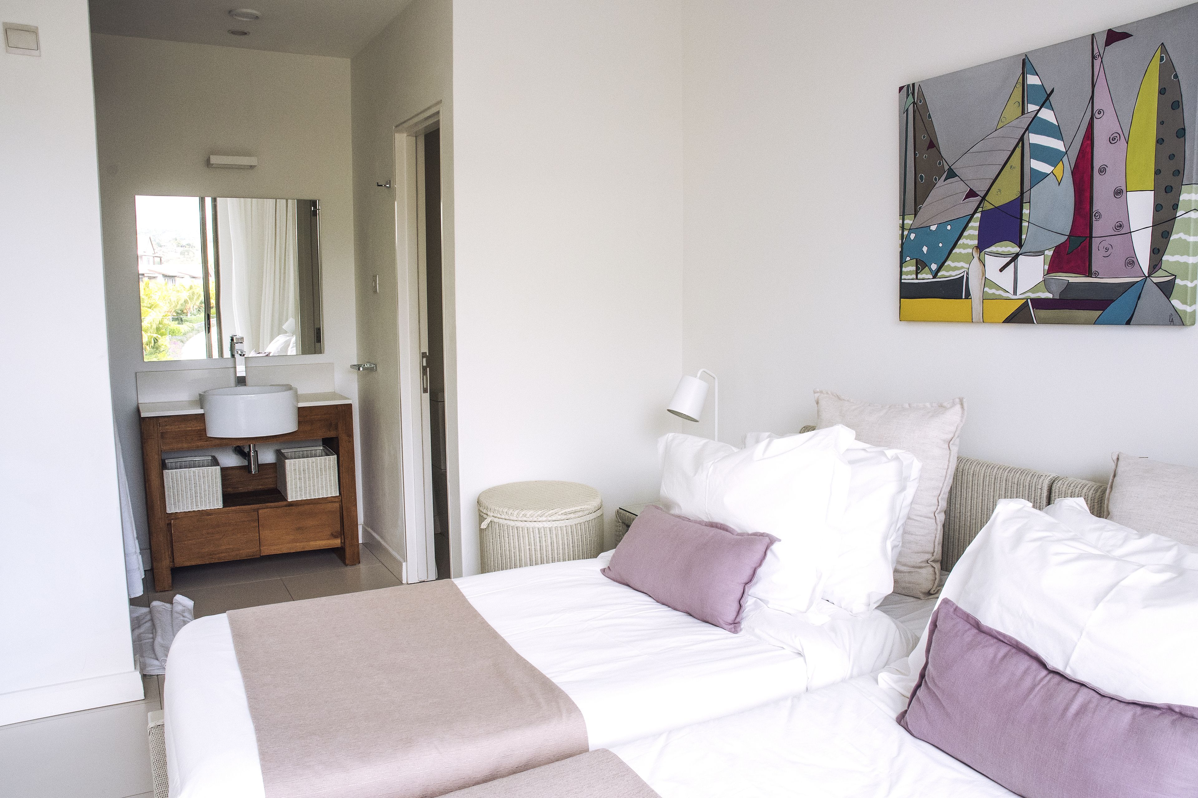 Een slaapkamer met een bed, kussens, een nachtkastje, een lamp, een schilderij, en een badkamer met een wastafel en een spiegel.