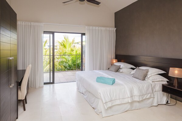 A modern bedroom with a large bed, a desk, and a balcony with a view of greenery.
