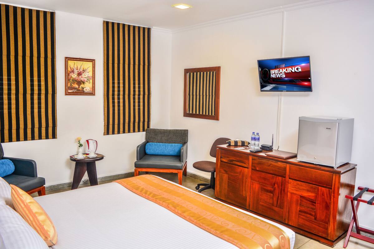 A hotel room with a large bed, a desk with a television, a chair, and a small table with a kettle and a vase.