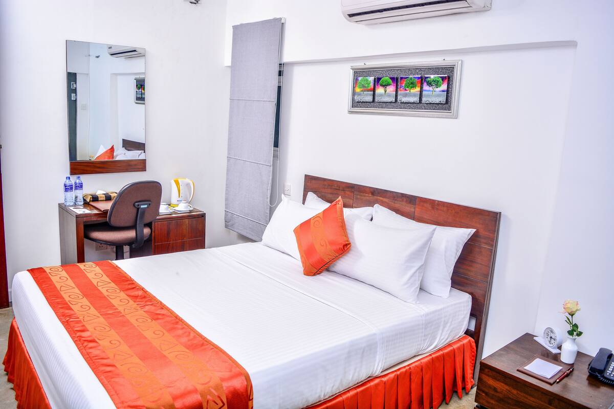A neatly arranged hotel room with a large bed, a desk with a chair, a small table with a vase, and a wall-mounted air conditioner.