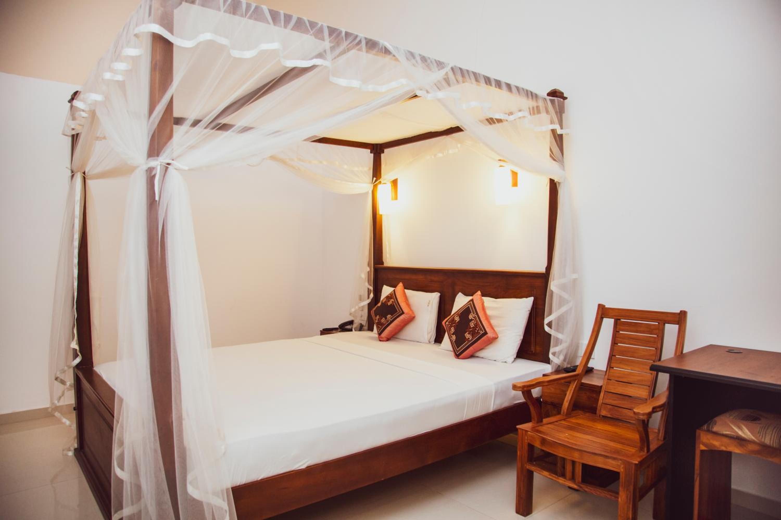 Una cama con dosel sobre cuatro columnas, ropa de cama blanca y almohadas decorativas, una mesita de noche de madera y una silla de madera.
