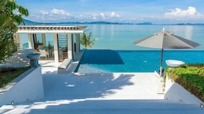 Un espace piscine avec vue sur la mer, un coin salon couvert et un escalier menant à la piscine.