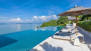 A infinity pool with a clear view of the ocean and distant islands, surrounded by lush greenery and white lounge chairs with umbrellas.